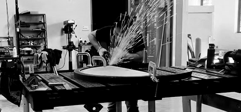 Hands-on metal fabrication, with sparks above a curved steel frame clamped to an assembly table